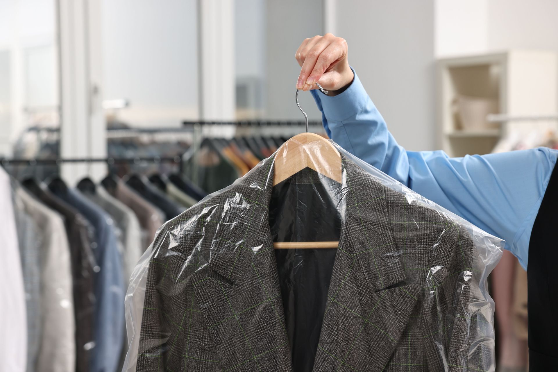 A person in a blue shirt holds a dark textured blazer in a clear plastic garment bag at a dry cleaner.