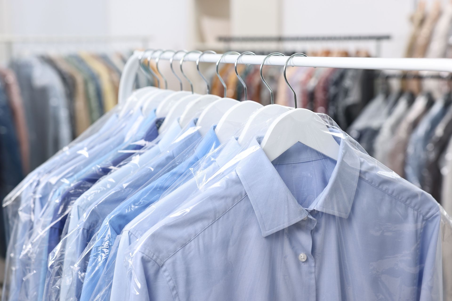 A rack of blue dress shirts on white hangers, encased in clear plastic garment covers, in a dry cleaner or clothing shop.