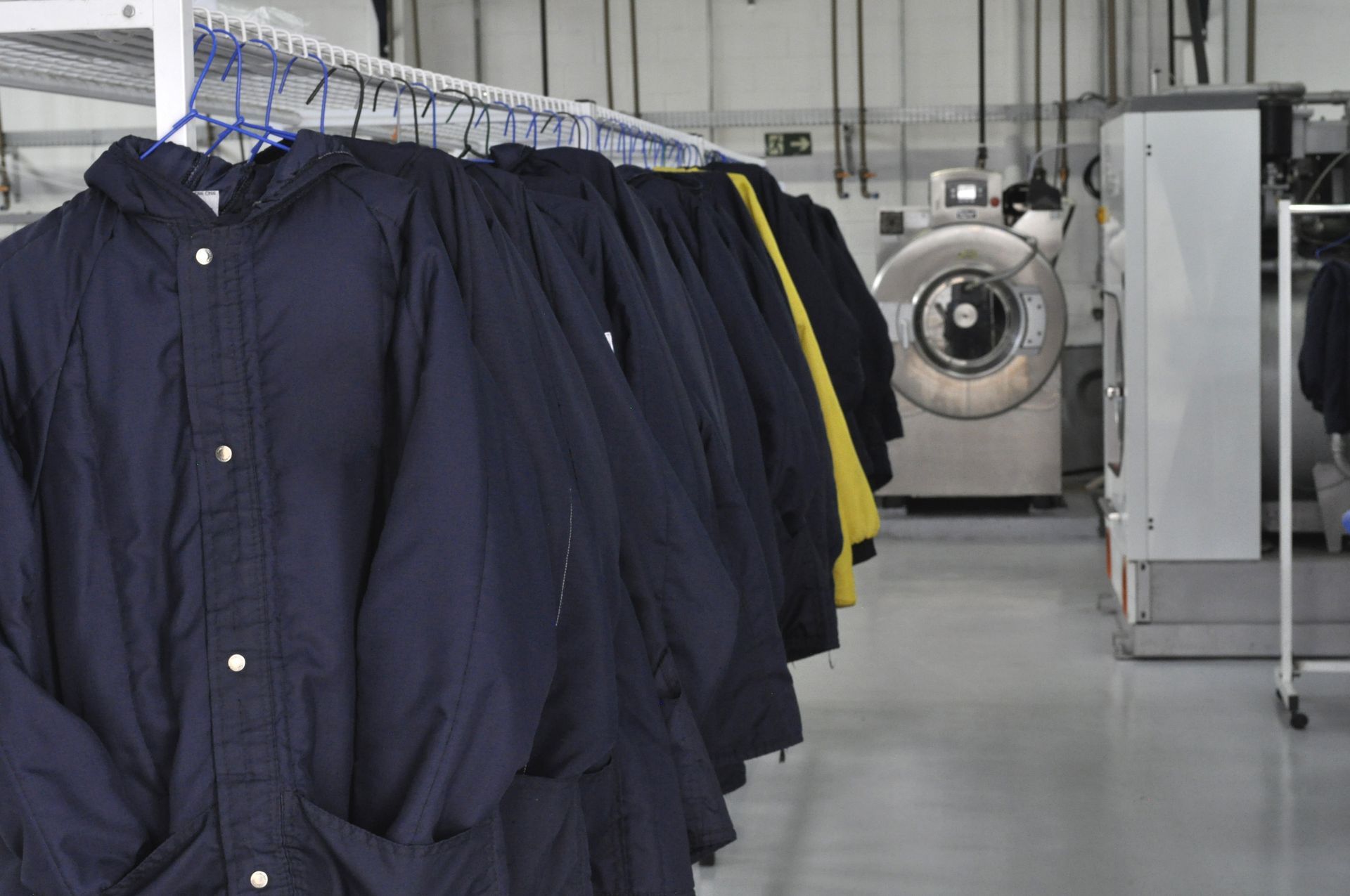 A rack of dark blue jackets hanging in a clean industrial laundry facility with large washing machinery in the background.