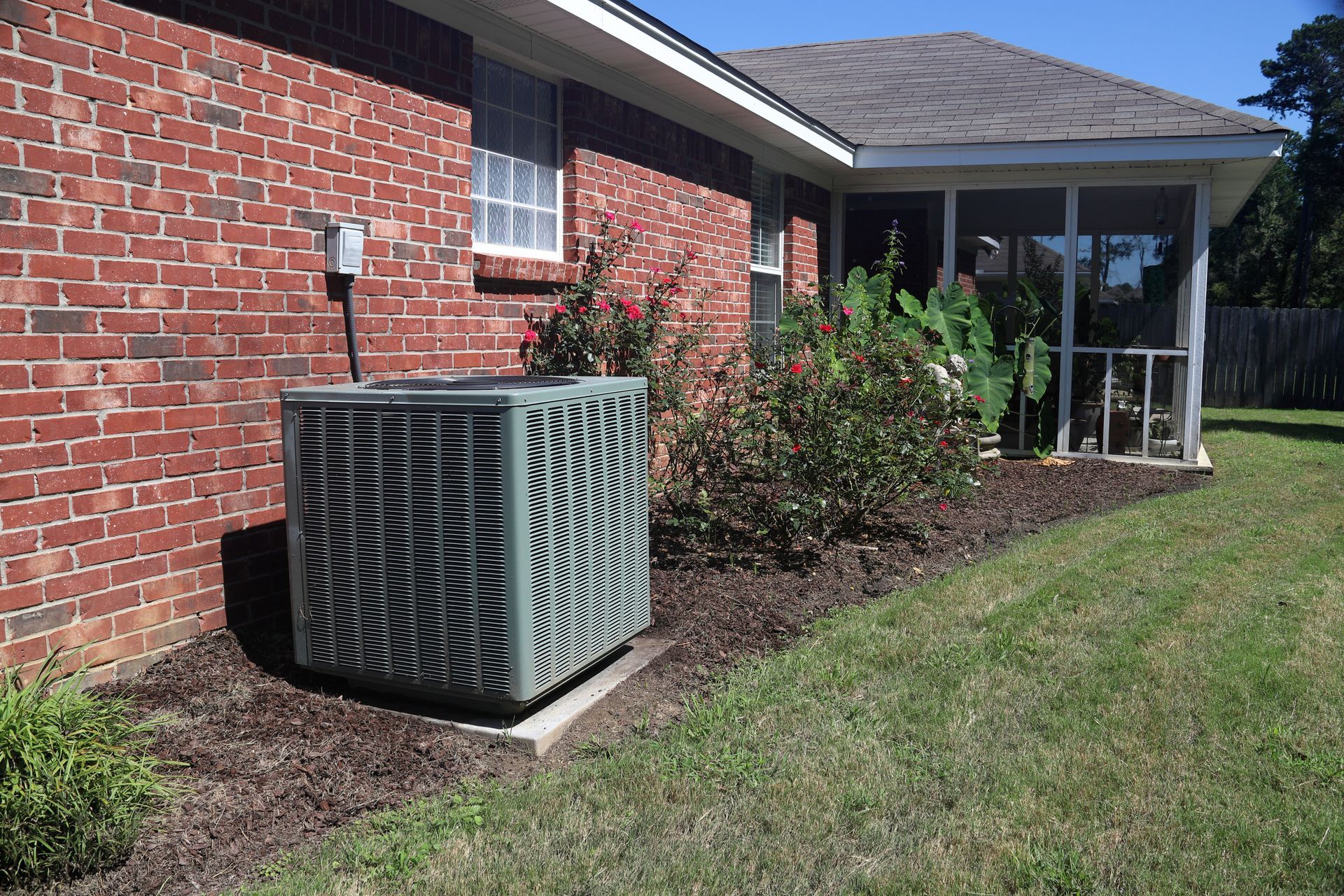 An outdoor HVAC condenser unit sits on a concrete pad next to a red brick house with a screened porch and garden beds.