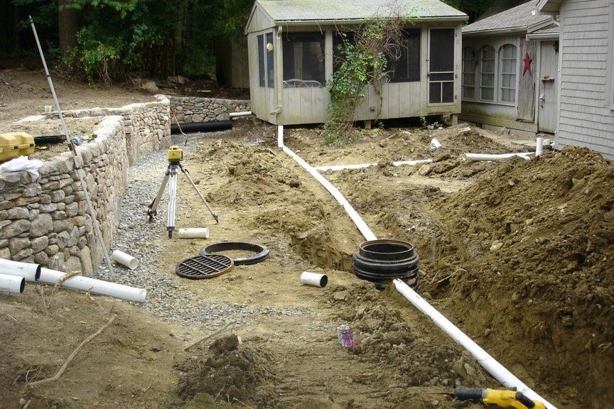 Construction site with white drainage pipes, a stone retaining wall, and a surveyor’s tripod in a backyard.