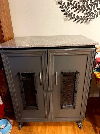 A gray two-door cabinet on wheels with metal mesh panels and a light granite-style countertop, set on a hardwood floor.