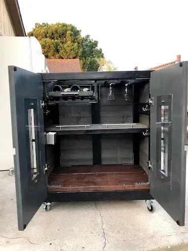 A black, mobile bar cabinet with glass-paneled doors, glass racks, and a wooden base standing on a concrete patio.
