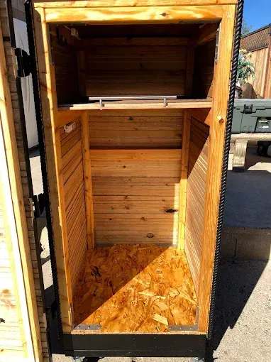 An open, upright wooden storage cabinet with a top shelf and a light-colored OSB wood floor, set outdoors.