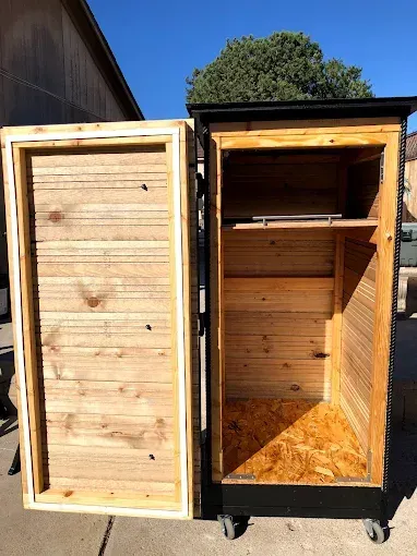 A wooden, wheeled storage cabinet stands outdoors with its door open, revealing a light-colored wood interior and one shelf.