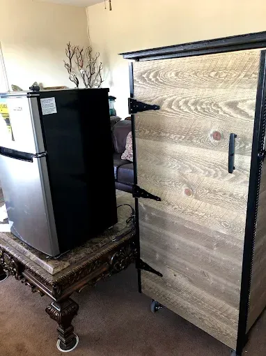 A mini-fridge sits on an ornate carved wooden table next to a tall, wood-paneled cabinet with black metal trim and hinges.