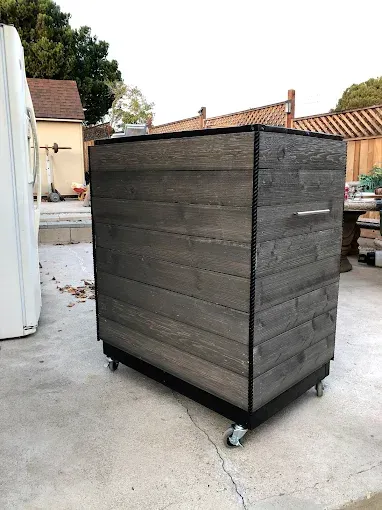 A gray, horizontal-planked wooden storage cabinet with a metal handle, positioned on casters on a concrete patio.