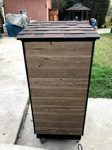A tall, wooden rectangular enclosure on wheels with a shingled, sloped roof, standing outdoors on a concrete patio.