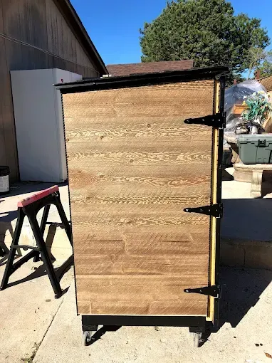A rectangular wooden cabinet on wheels with black hinges and trim, standing outdoors on a concrete patio.