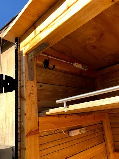 Interior view of a small wooden structure featuring a metal L-bracket at the corner, two light strips, and a shelf.