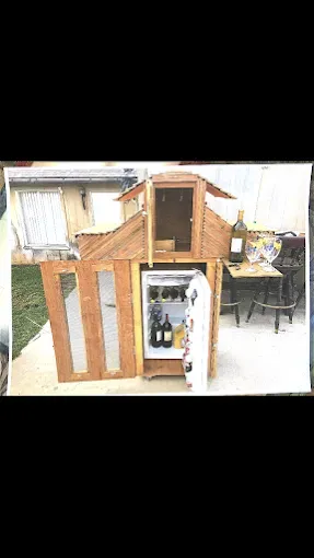 A wooden structure resembling a small house built around a mini-fridge stocked with wine bottles, outdoors on a patio.