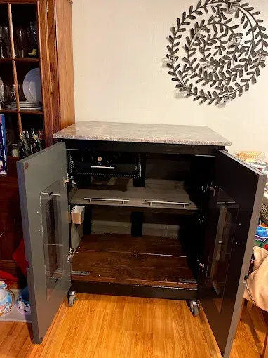 A black, wheeled kitchen island cabinet with an open door, a stone top, and two interior shelves on a wooden floor.
