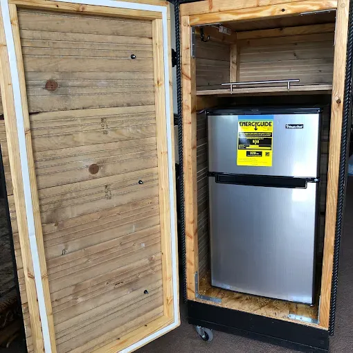 A silver mini-fridge sits inside a custom wooden cabinet with a hinged door, mounted on small wheels.