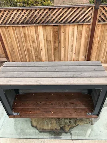 A wooden outdoor bench with a light-gray slatted top and a dark-brown wooden shelf base, set against a wooden fence.