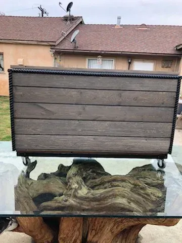 A wooden, plank-style chest with rope trim and casters sits atop a glass-topped driftwood coffee table outdoors.