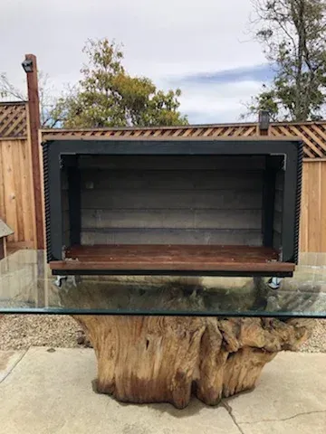 A rectangular, open-front wooden cabinet with black metal trim sits on top of a glass table supported by a tree stump base.