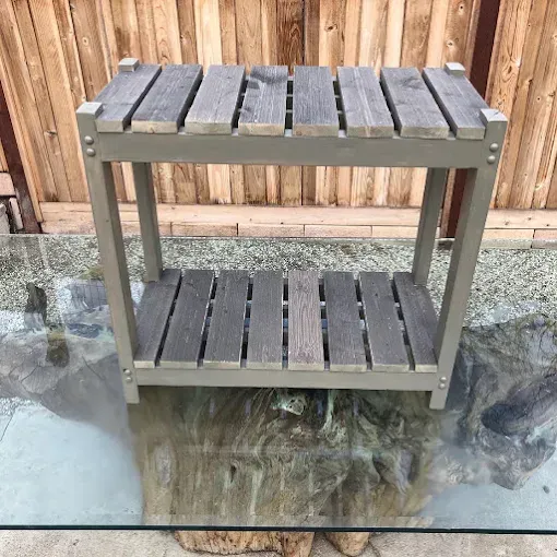 A small, two-tiered wooden plant stand with gray-painted legs and weathered, horizontal slat shelves on a glass table.