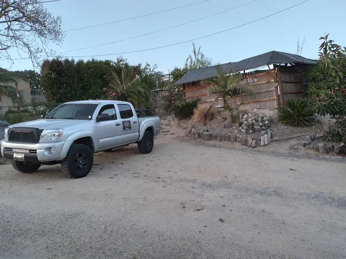 A silver pickup truck parked on a dirt lot next to a rustic, partially fenced structure under a clear sky.