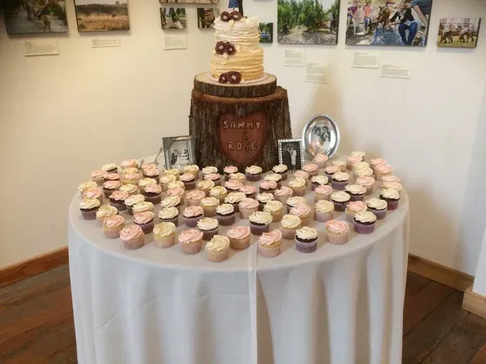A wedding cake on a rustic log stand surrounded by many cupcakes on a round white table in an art gallery.