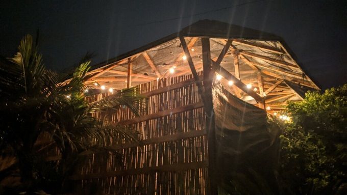 A wooden structure with a slatted wall illuminated by string lights against a dark night sky, flanked by palm leaves.