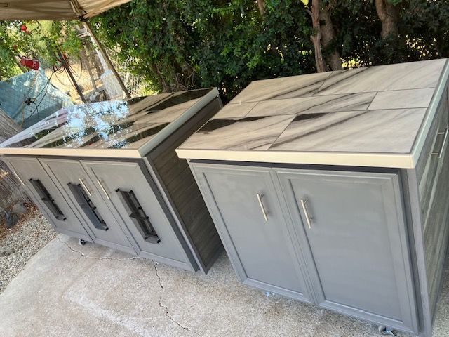Two outdoor kitchen cabinets on casters with grey doors and light-colored tiled countertops sitting on a patio.