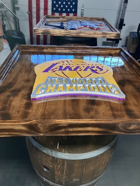 A wooden Lakers championship sign encased in resin, mounted on a barrel in a workshop with an American flag background.