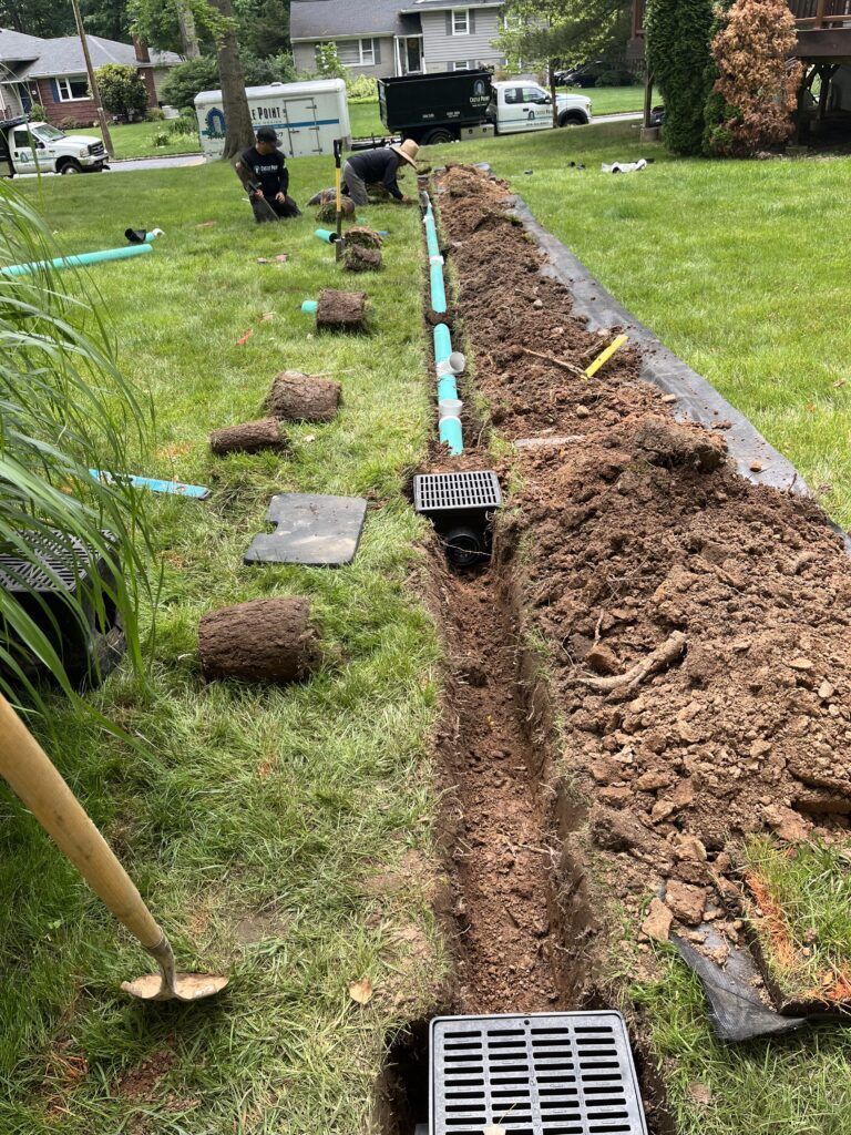 Workers install a blue drainage pipe and grate systems in a trench through a residential lawn.