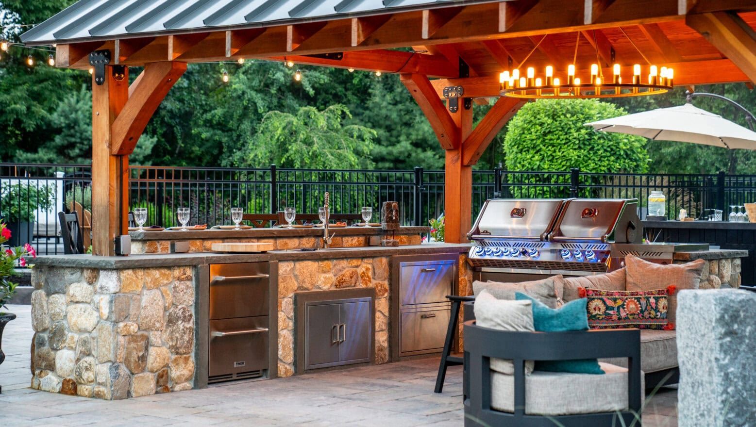 Outdoor kitchen under a wooden pavilion featuring stone counters, a stainless steel grill, and lounge seating.