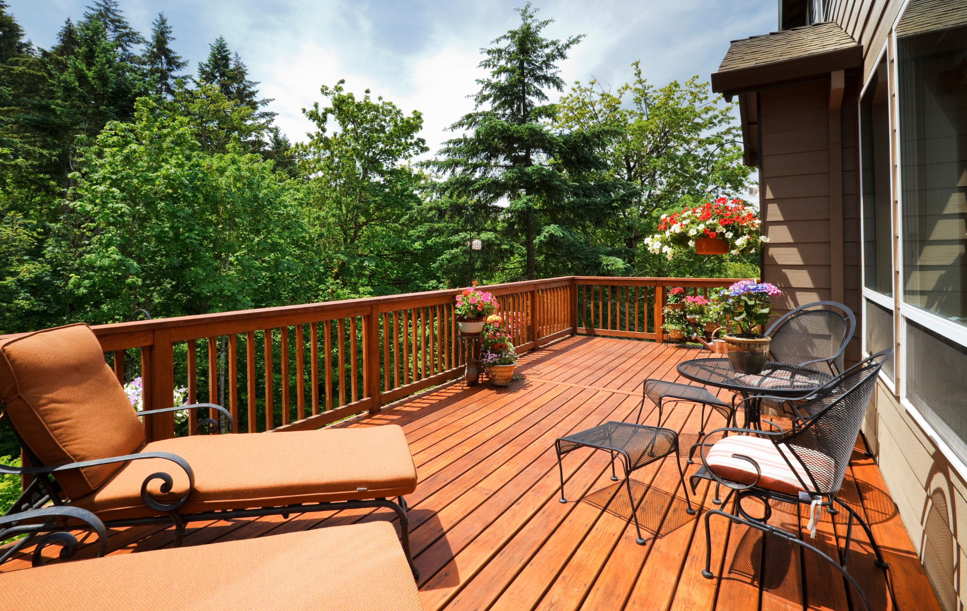 A wooden deck with a lounge chair, a table and chairs, and potted flowers, overlooking a forest under a cloudy sky.