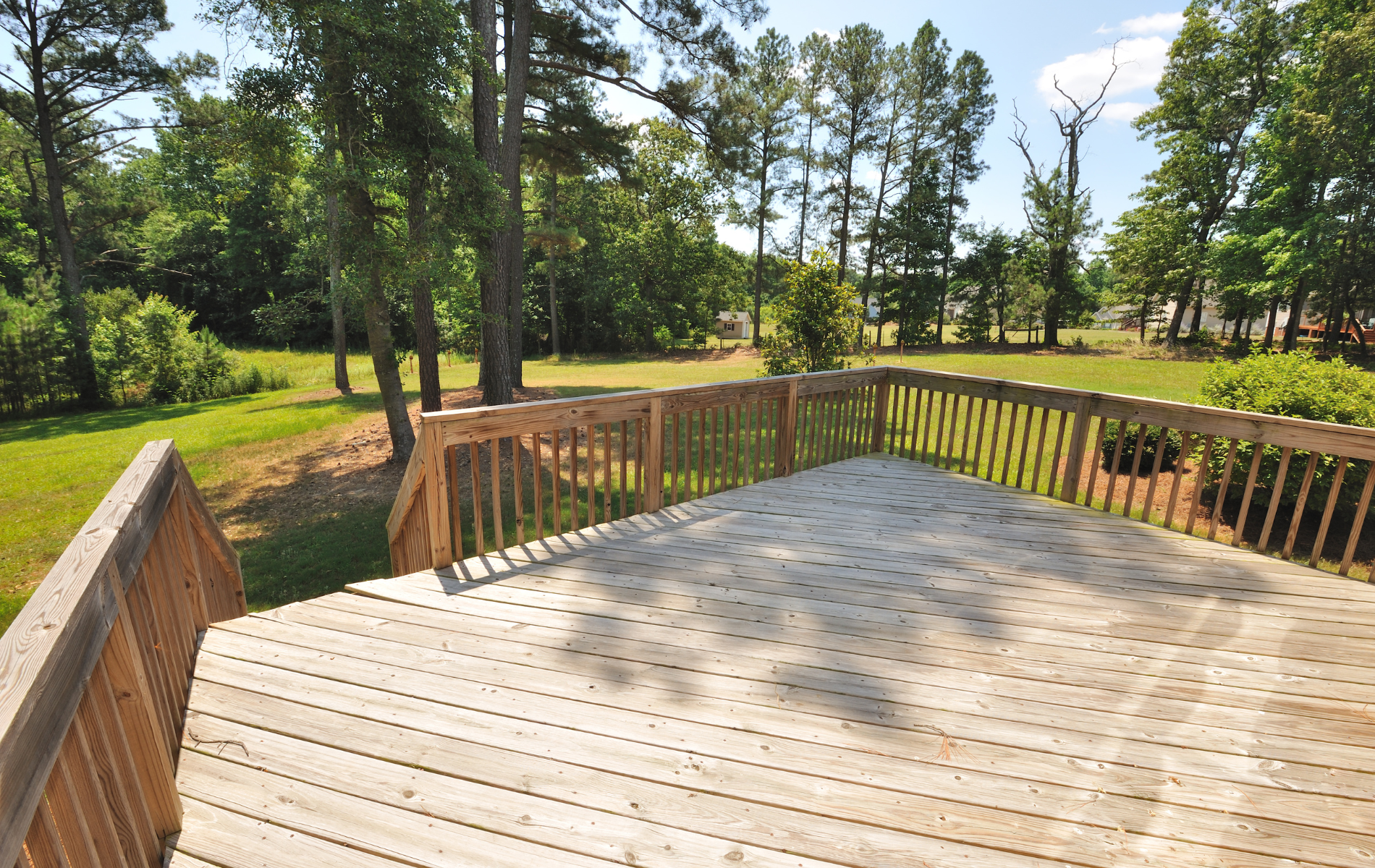 An elevated wooden deck with railing overlooks a grassy backyard filled with mature trees under a blue sky.