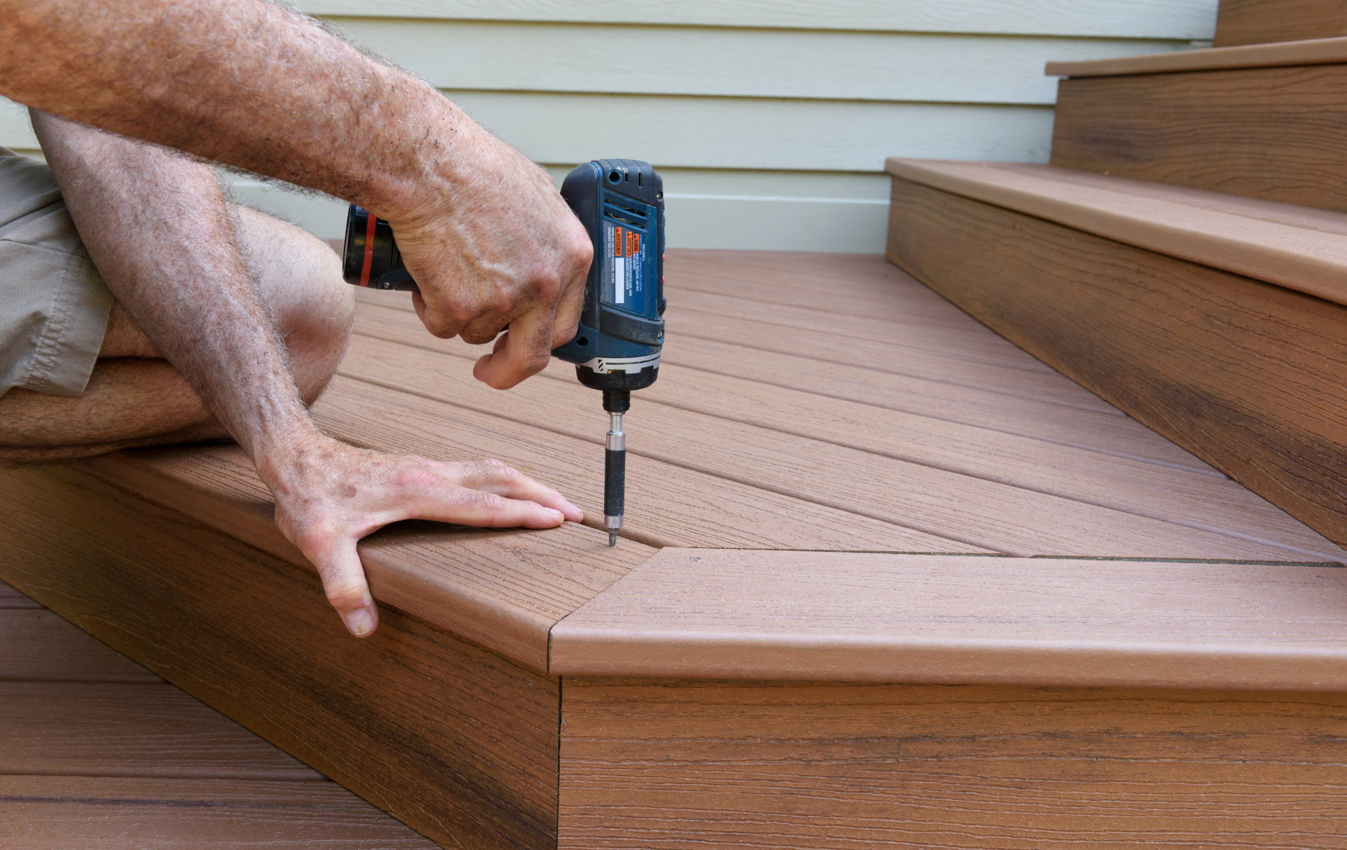 A person uses a power drill to fasten a brown composite deck board to stairs on an outdoor deck.