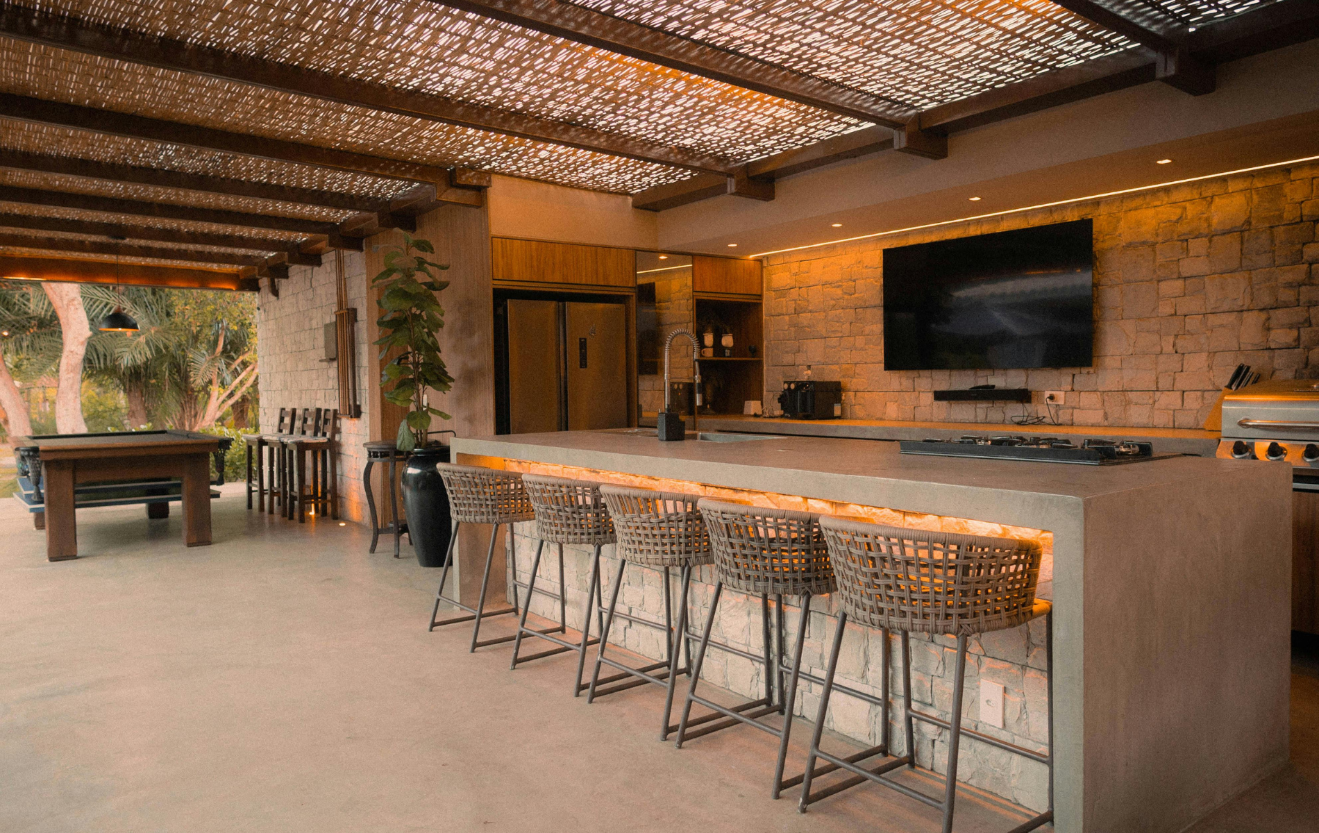 A modern outdoor kitchen with a concrete bar, woven stools, a wall-mounted TV, and a covered patio area.