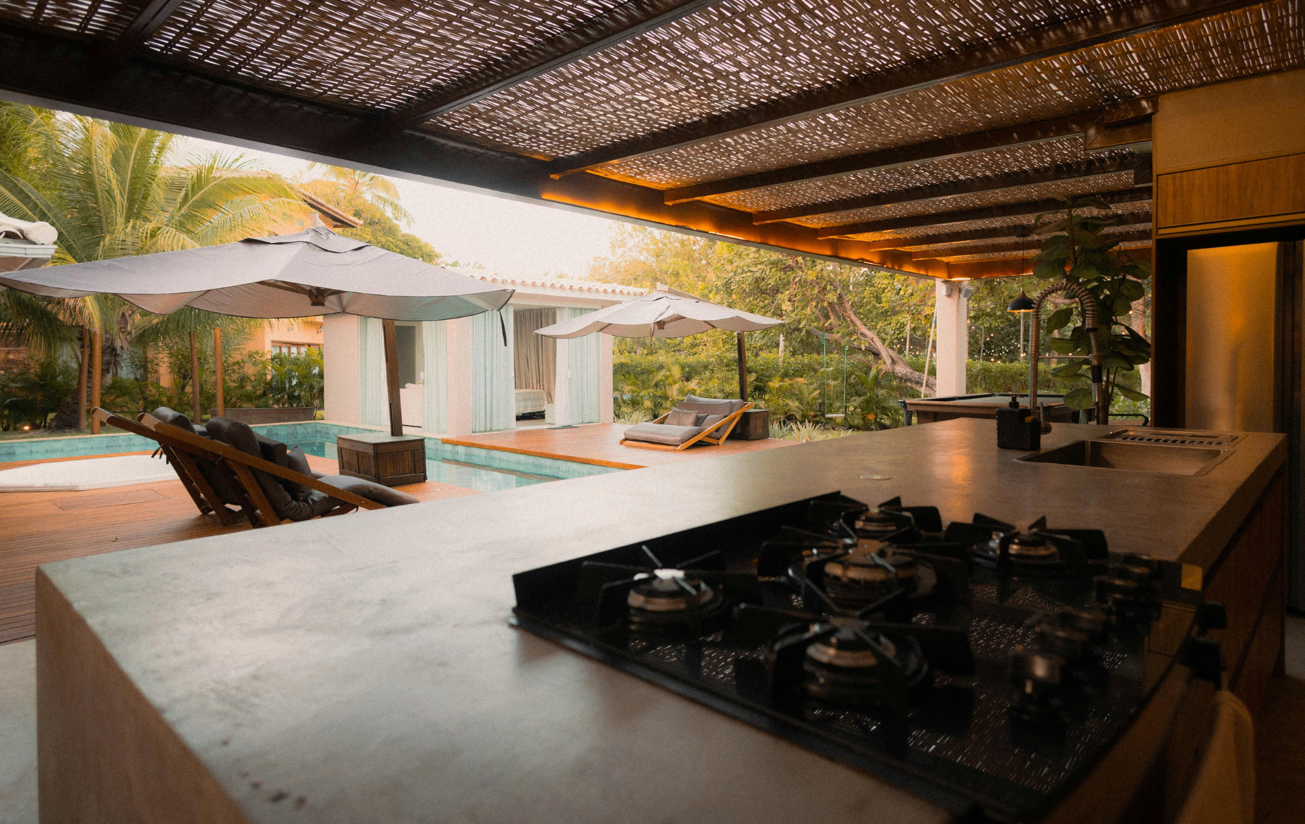 A sleek outdoor kitchen with a stovetop overlooks a sunlit swimming pool area with lounge chairs and umbrellas.