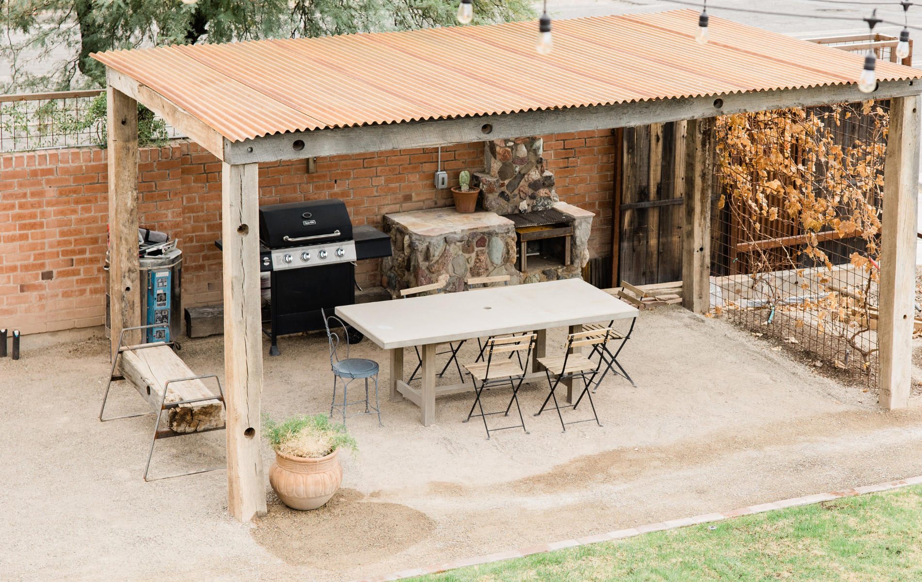 An outdoor dining area under a rustic corrugated metal roof with a grill, stone countertop, table, and chairs.