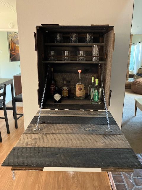 A wall-mounted, rustic wooden bar cabinet with an open drop-down door, stocked with glass tumblers and liquor bottles.