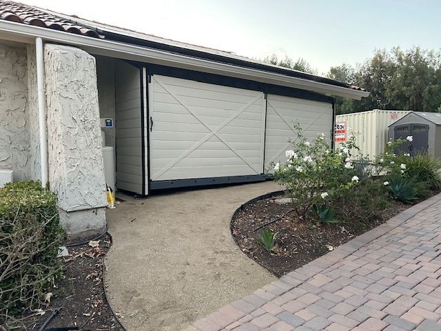 A paved path leads to a large, light-colored sliding barn door attached to the side of a house with nearby landscaping.