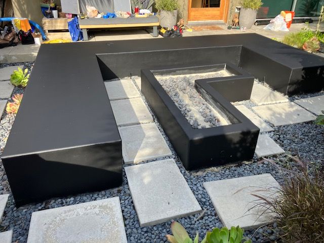 A black, L-shaped modern outdoor seating area surrounding a central recessed fire pit set amidst square stone pavers.