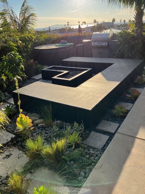 A modern, L-shaped black fire pit sits on a patio next to a grill, surrounded by landscaped plants at sunset.