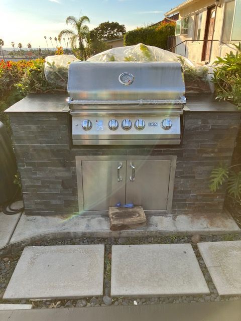 A built-in stainless steel grill with a stone facade, situated on a patio outside a house.