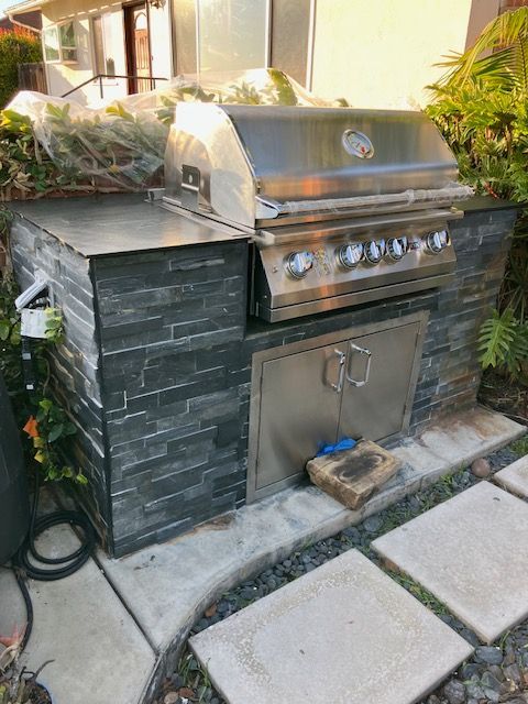 A stainless steel built-in barbecue grill with gray stone siding, set on a concrete patio in a residential backyard.