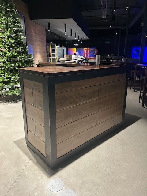 A rectangular wooden bar with a dark metal frame stands in a dimly lit, industrial-style room with a Christmas tree nearby.
