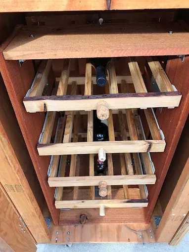 A rustic wooden wine rack with four pull-out shelves holding three wine bottles inside an open cabinet.