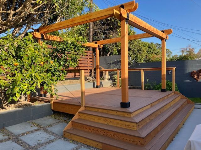 A wooden deck with a built-in pergola and wide front steps in a sunny backyard garden.