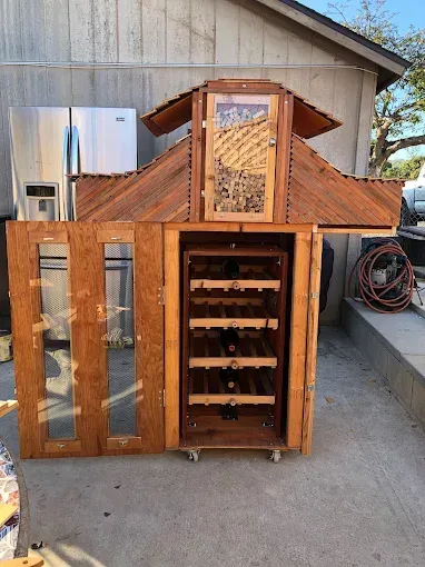 A custom-built wooden wine cabinet on wheels, featuring glass-paneled doors, shelving, and a decorative roof structure.