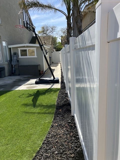 A side yard with a white vinyl fence, a strip of dark gravel, an artificial grass lawn, and a portable basketball hoop.