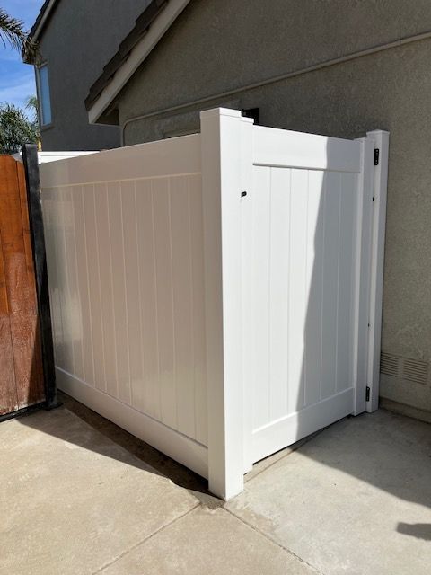 A white vinyl privacy screen with a gate stands on a concrete patio next to a tan exterior house wall.