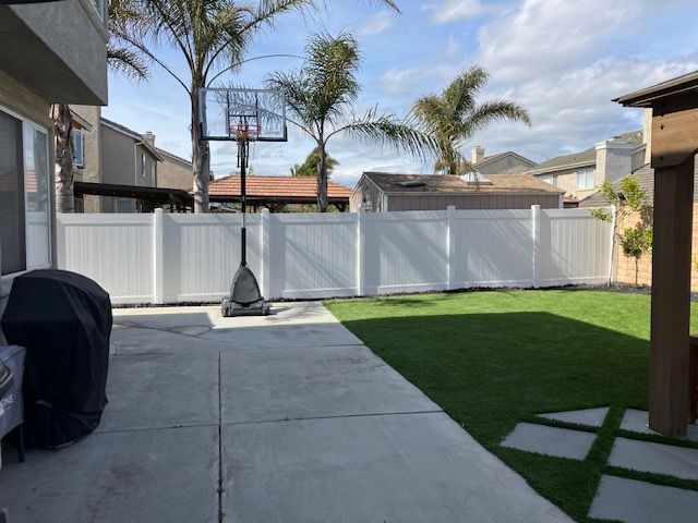 A backyard with a concrete patio, a covered grill, a basketball hoop, white vinyl fencing, and a patch of green lawn.