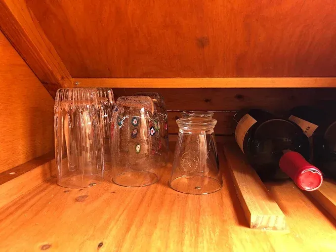 Several clear glasses sit on a wooden shelf next to two dark wine bottles lying on their sides.