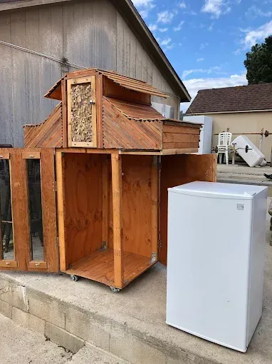 A wooden, multi-level animal enclosure with a decorative door sits outdoors next to a small, white refrigerator.