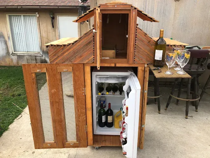 A custom wooden bar cabinet with a roof-like structure, an open door, a small refrigerator inside, and wine glasses nearby.